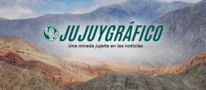 Portada de JUJUYGRÁFICO con paisaje montañoso de la Quebrada de Humahuaca en tonos naturales y cielo parcialmente nublado. En el centro destaca el logo con un cactus o cardón estilizado y el nombre “JUJUYGRÁFICO” en tipografía grande color verde azulado, acompañado del eslogan “Una mirada jujeña en las noticias”. Diseño panorámico realista optimizado para visualización en computadoras y dispositivos móviles. Jujuy Gráfico noticias de Jujuy